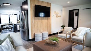 A modern living room with a sofa, armchairs, a coffee table, a TV mounted on the wall, and a kitchen area in the background.