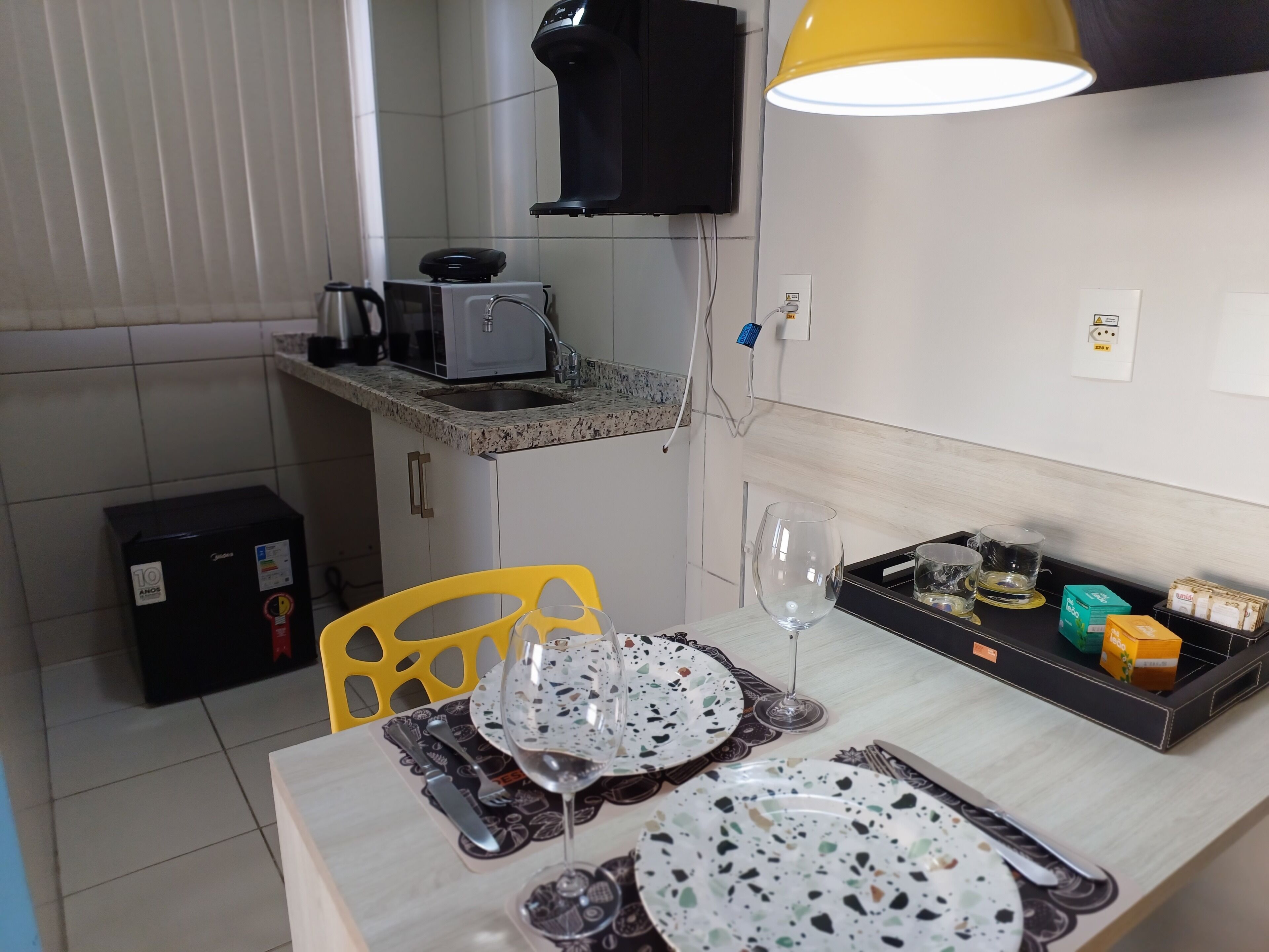 Uma cozinha compacta com micro-ondas, cafeteira e uma pequena geladeira. Uma mesa de jantar com pratos, talheres e uma taça de vinho está posta.