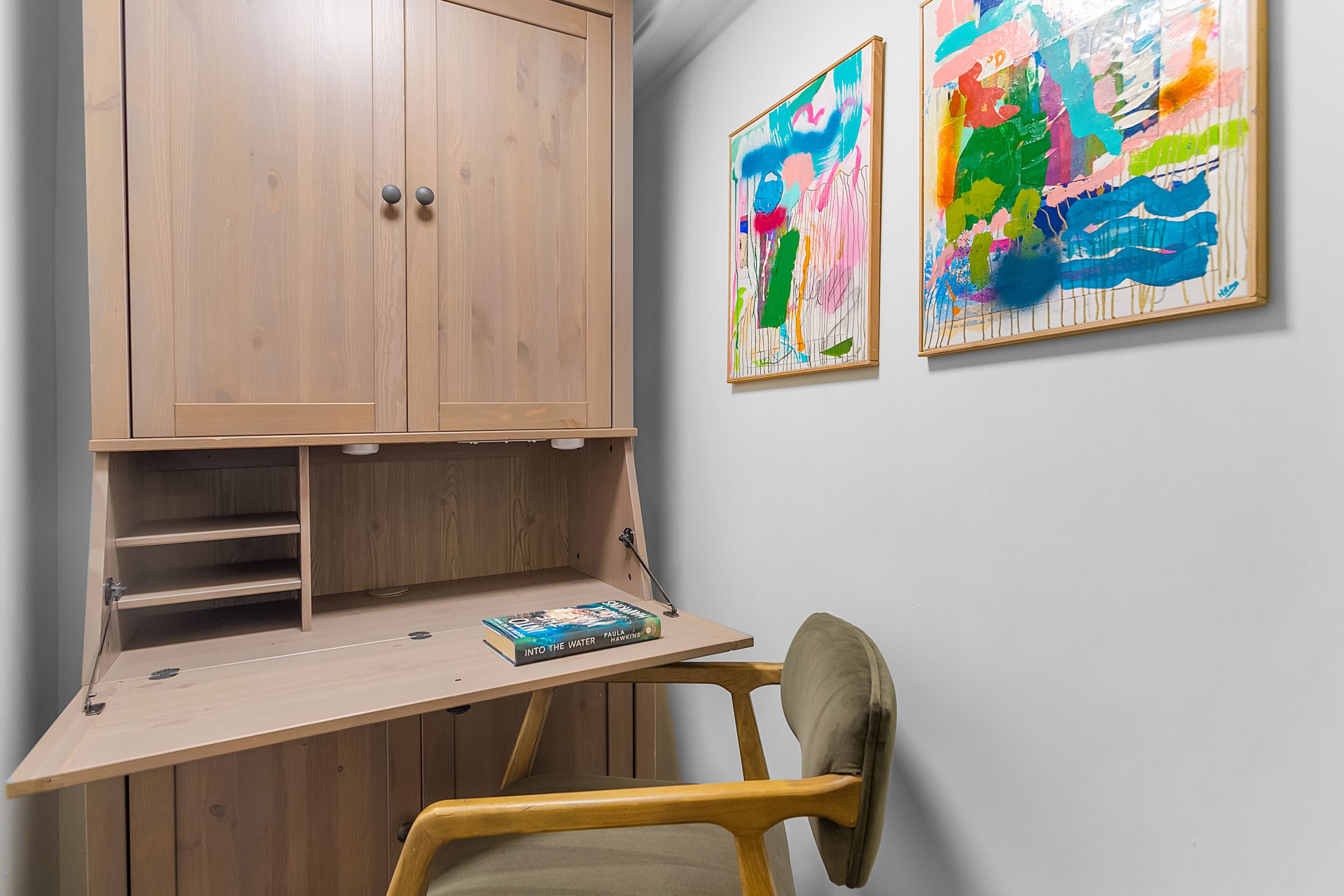 Ein Zimmer mit einem Holztisch, einem Stuhl, einem Bücherregal und zwei abstrakten Bildern an der Wand.
