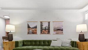 A modern living room with a green sofa, wooden coffee table, and two poufs. There are two lamps on side tables and three framed pictures on the wall. - Lilac in Law (Seattle)