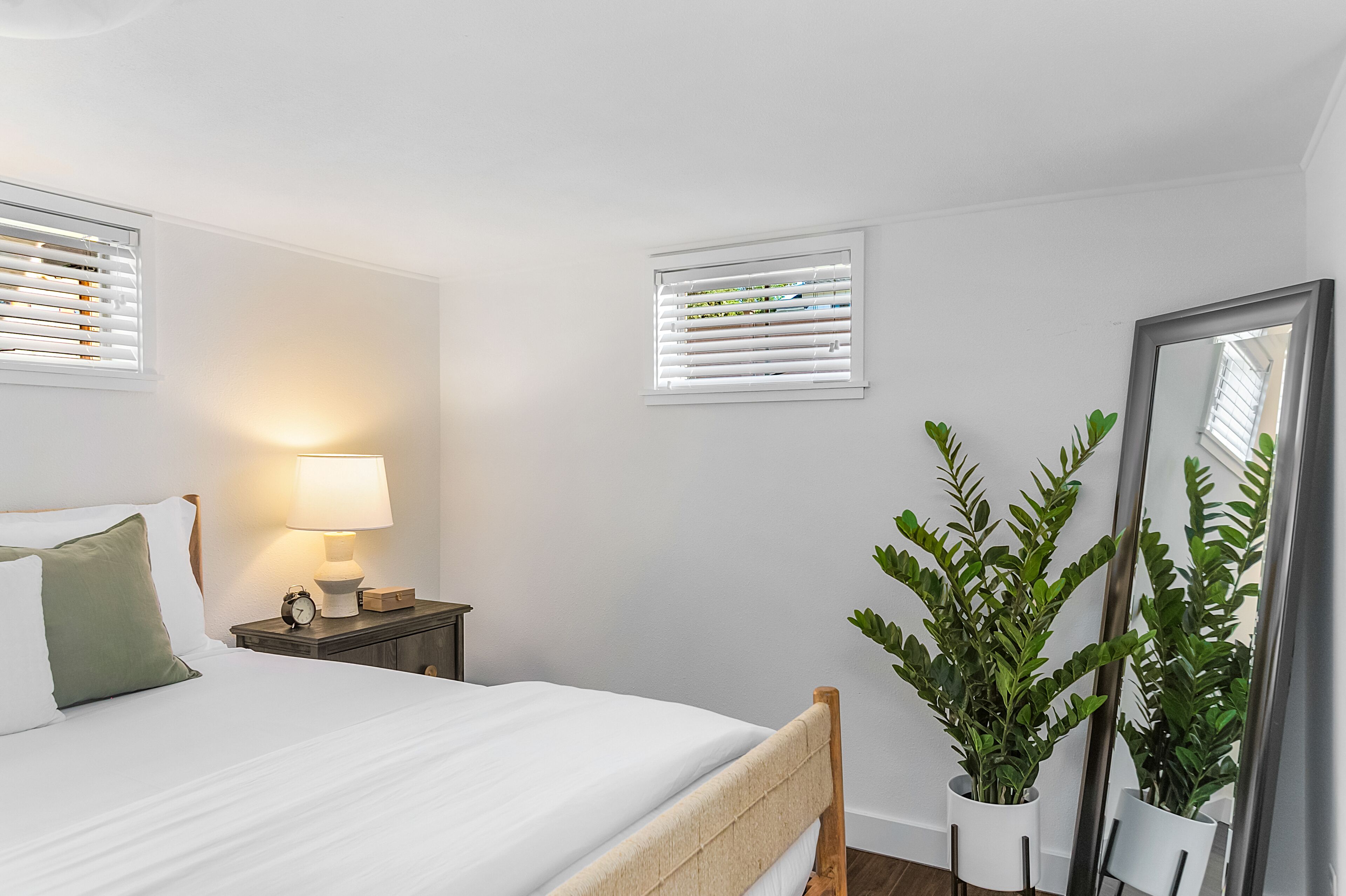 A neatly arranged bedroom with a bed, bedside lamp, two windows with blinds, a nightstand, a mirror, and a potted plant.