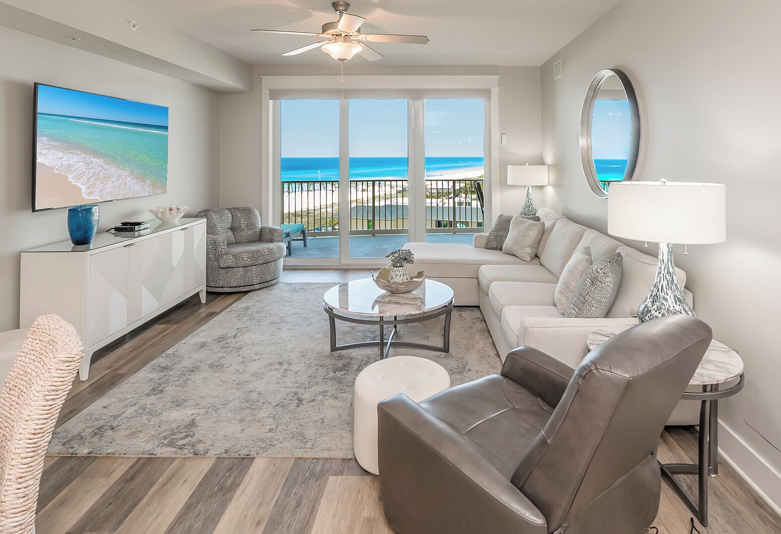 A spacious living room with a large TV, a ceiling fan, and a view of the beach through the sliding glass doors.