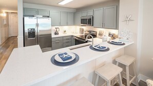 A modern kitchen with grey cabinets, a stainless steel refrigerator, a built-in microwave, and a coffee machine. There are three bar stools and a white countertop with blue placemats. - LAST Minute Cancellation! Luxurious gorgeous condo and includes beach service! (Panama City Beach)