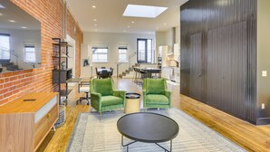 A spacious living area with a brick wall, green armchairs, a black coffee table, and a staircase leading to a kitchen.