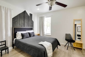A modern bedroom with a large bed, a ceiling fan, a wooden headboard, a bedside table, a chair, and a mirror.