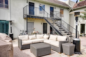 A covered outdoor seating area with a sectional sofa and ottomans, a staircase leading to a balcony, and a trash can.