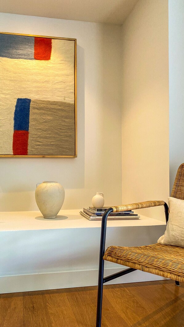 A modern living room with a white console table, a wicker chair, a white vase, and a painting with red and blue stripes.