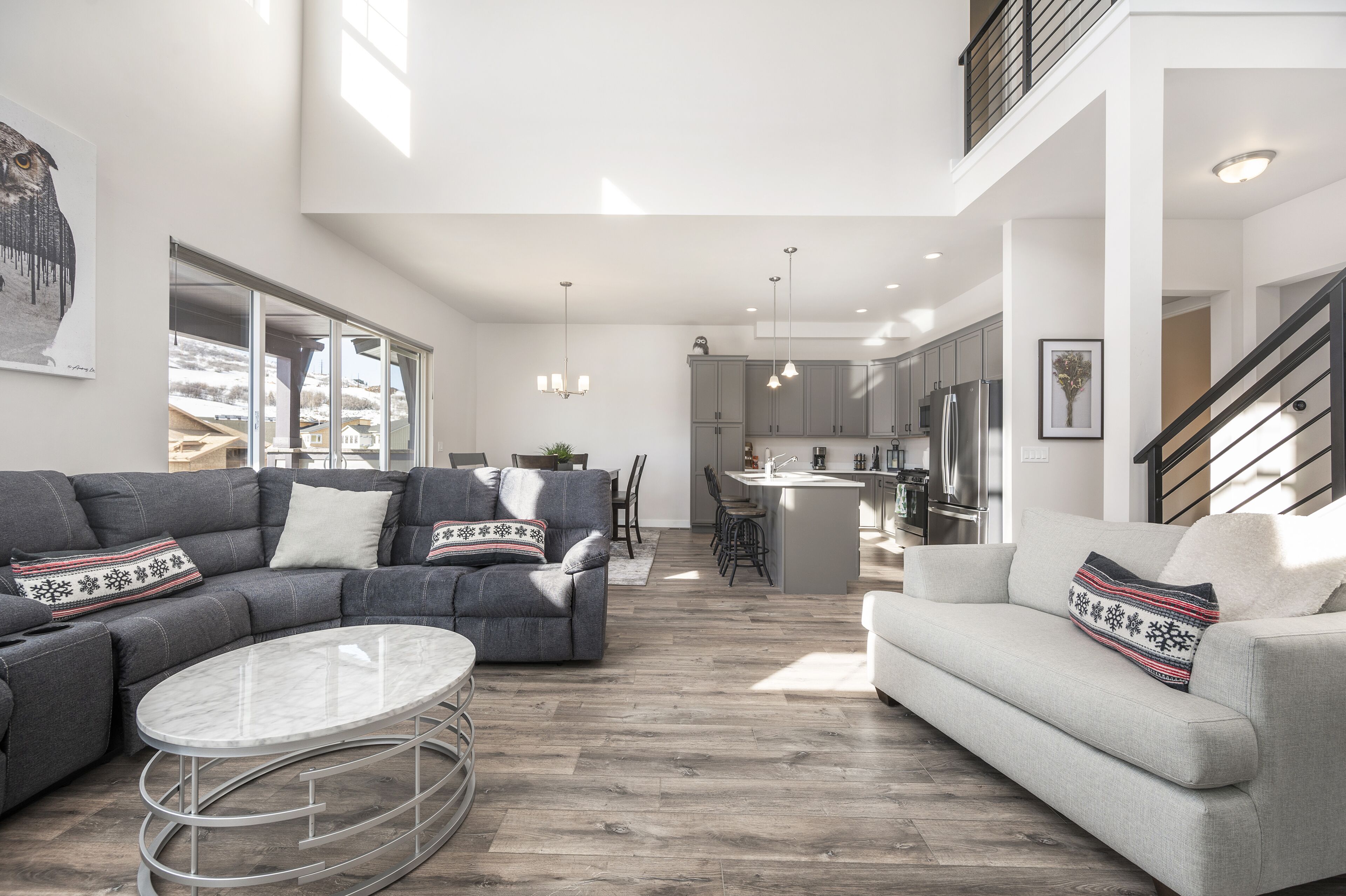 A spacious living area with a gray sectional sofa, a round coffee table, and a gray sofa. A kitchen with modern appliances and a dining area with chairs are visible in the background.