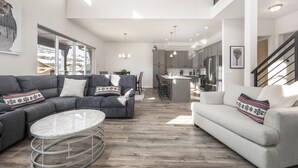 Een ruime woonkamer met een grijze modulaire bank, een ronde salontafel en nog een grijze bank. Achtergrond toont een keuken met moderne apparatuur en een eethoek met stoelen.