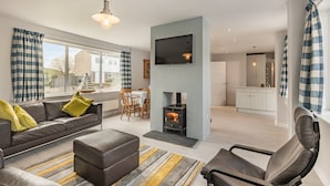 A modern living room with a sofa, ottoman, and a fireplace. There is a dining area with a table and chairs, and a kitchen in the background. - A stunning location for exploring the stunning North Norfolk Coast. (Brancaster Staithe, near King&rsquo;s Lynn)