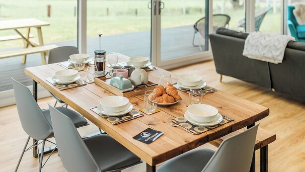 Una mesa de comedor dispuesta con platos, vasos y una cafetera, con vistas a un paisaje pintoresco.