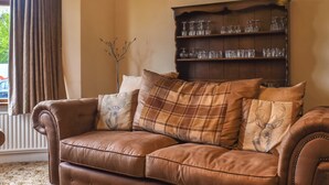 A brown leather sofa with plaid cushions, a wooden bookshelf with glassware, and a window with curtains. - 4 bedroom accommodation in Clitheroe (Clitheroe)