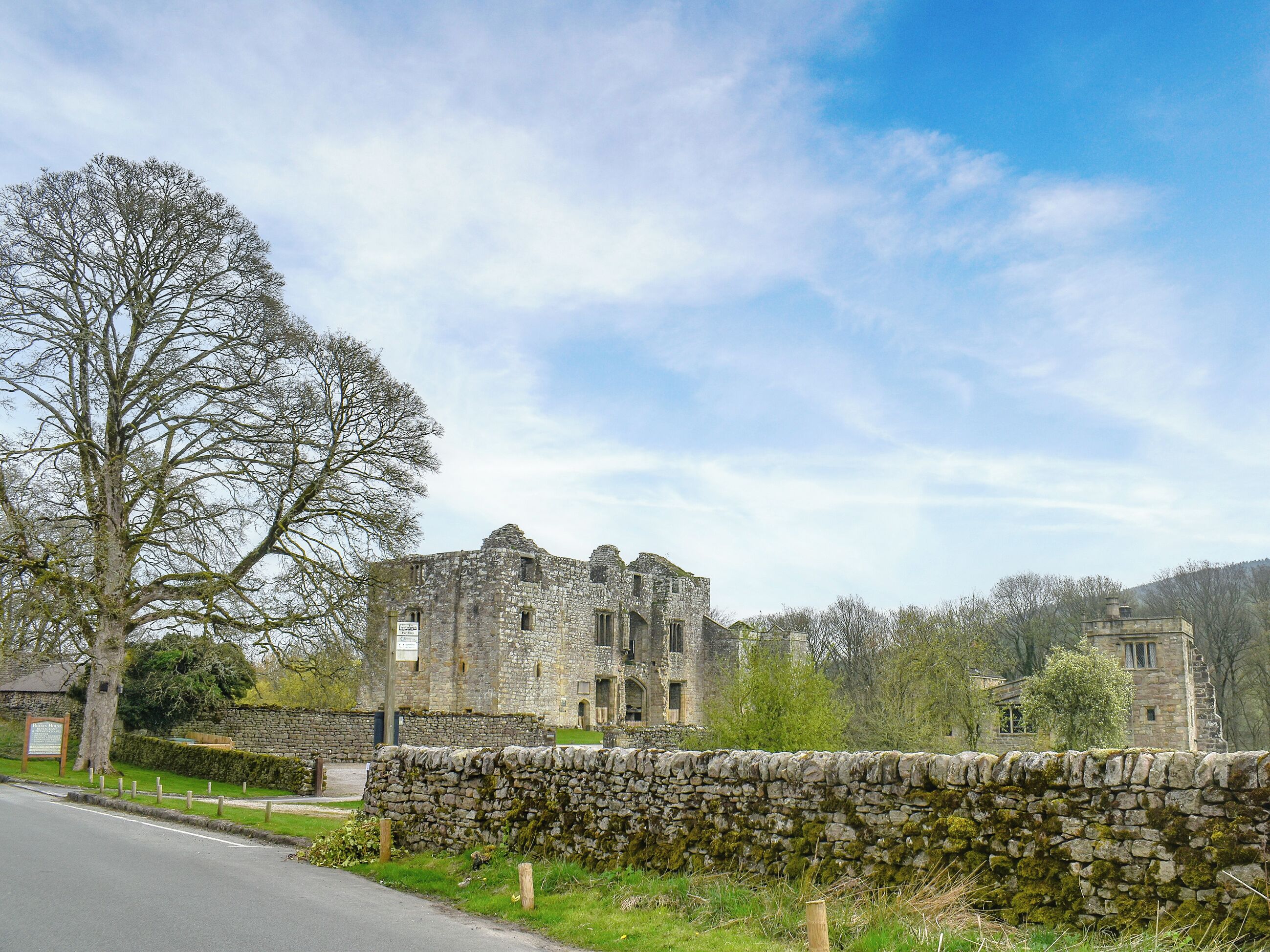 A historic stone building with a large tree in the foreground, a stone wall, and a clear sky.