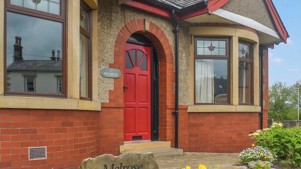 A red-brick house with a red door, a small garden with flowers, and a street sign reading "2 Melrose". - 4 bedroom accommodation in Clitheroe (Clitheroe)
