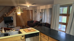 A compact kitchen with wooden cabinets, a dishwasher, and a sink. There is a dining area with a couch and a TV in the background. - The Big House in the Heart of Errol (Errol)
