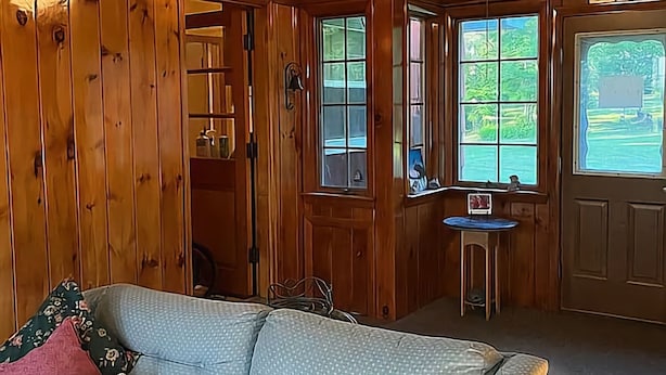 A cozy room with wooden paneling, a sofa with patterned cushions, a small table, and a window with a view of greenery. - The Big House in the Heart of Errol (Errol)