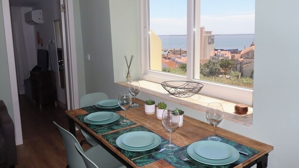 A dining area with a table set for four, a window with a view of buildings and a body of water, and a door leading to another room. - Lisbon, T2 renewed duplex with river view, Beato (Lisboa ciudad)