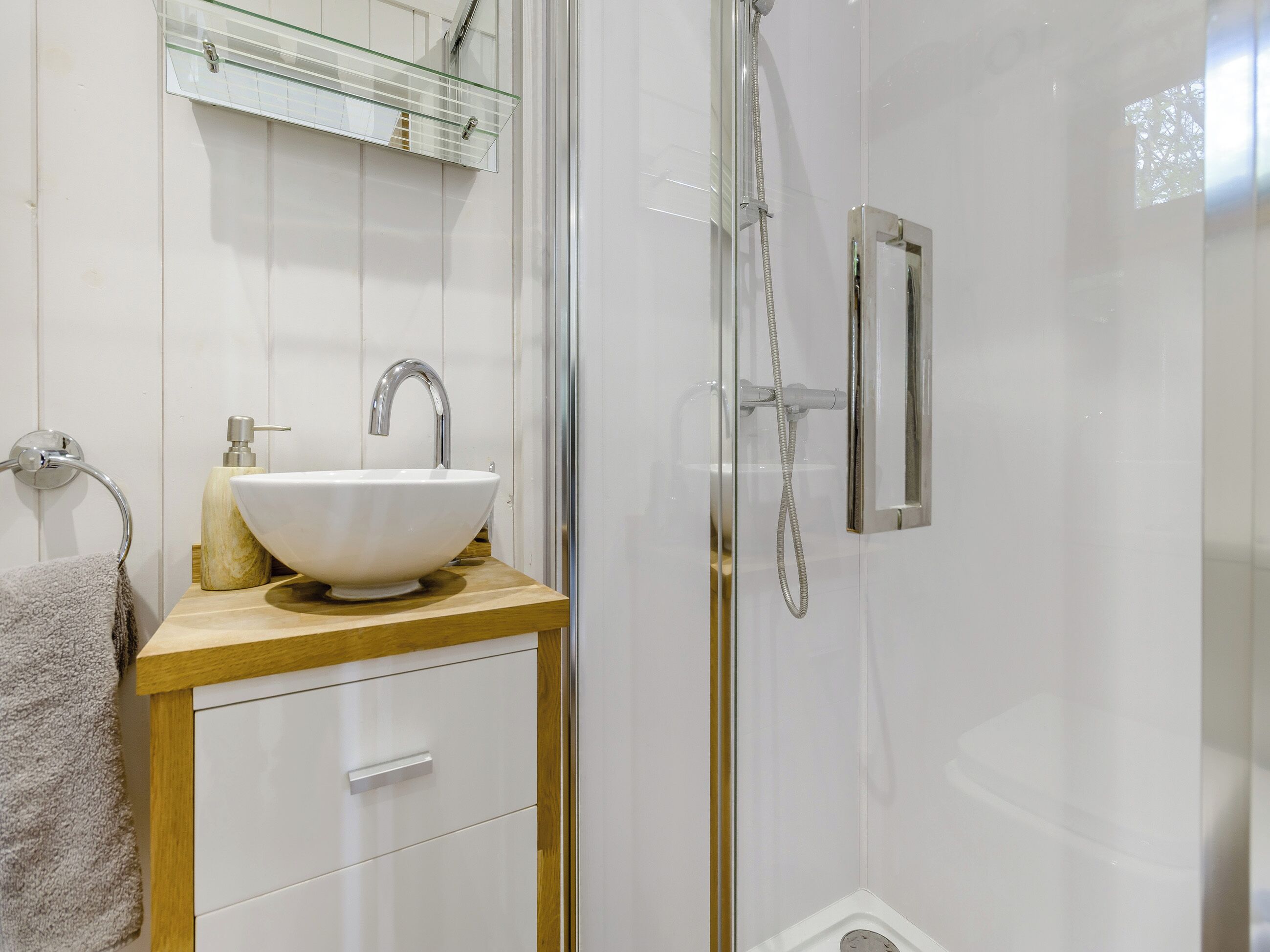 A bathroom with a glass shower enclosure, a white sink with a marble-like soap dispenser, and a wooden vanity with a towel rack.