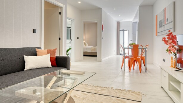 A modern living room with a glass coffee table, a grey sofa with cushions, a dining area with orange chairs, and a white sofa in the background.