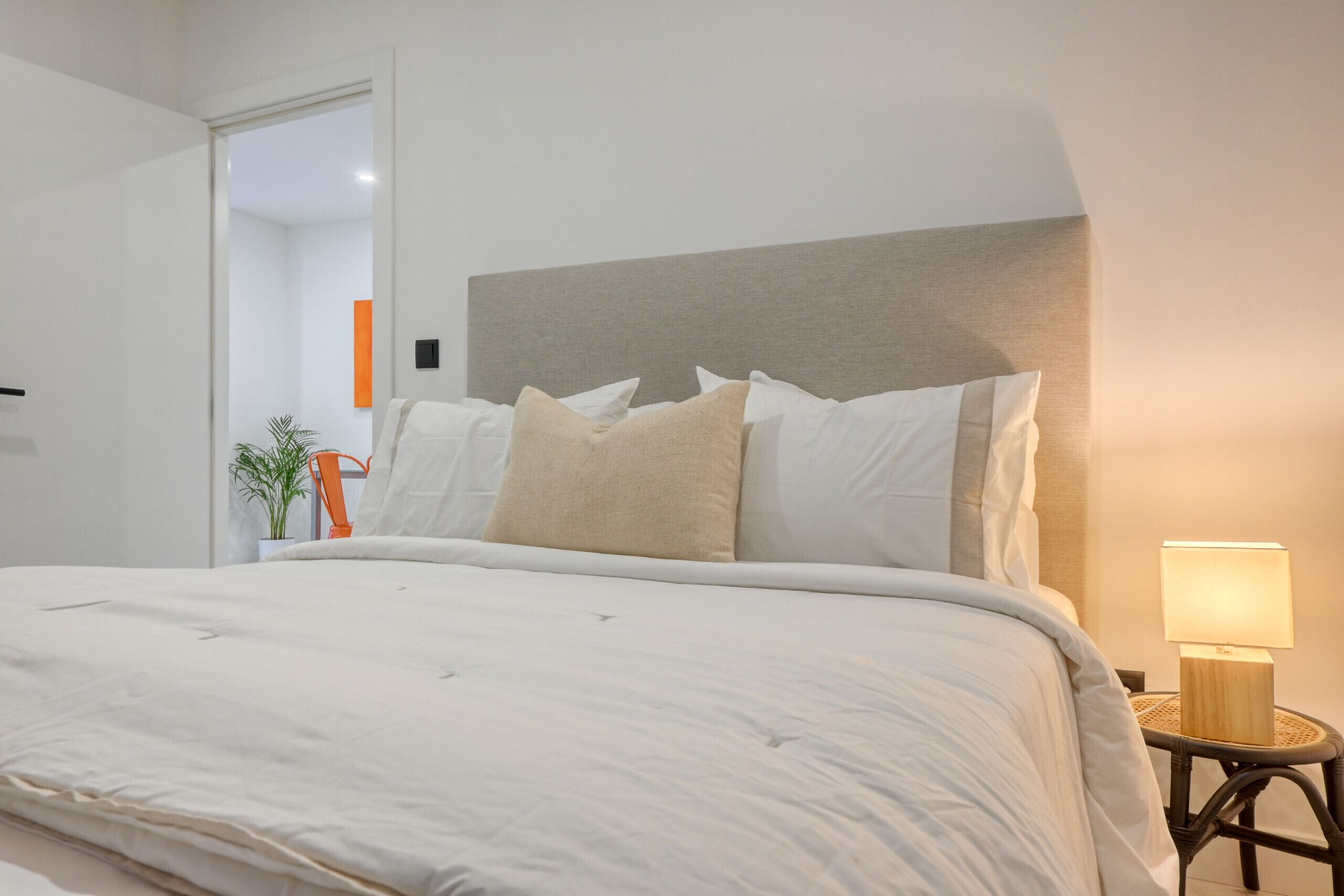 Una cama bien hecha con cabecera gris, ropa de cama blanca y una mesita de noche con lámpara.