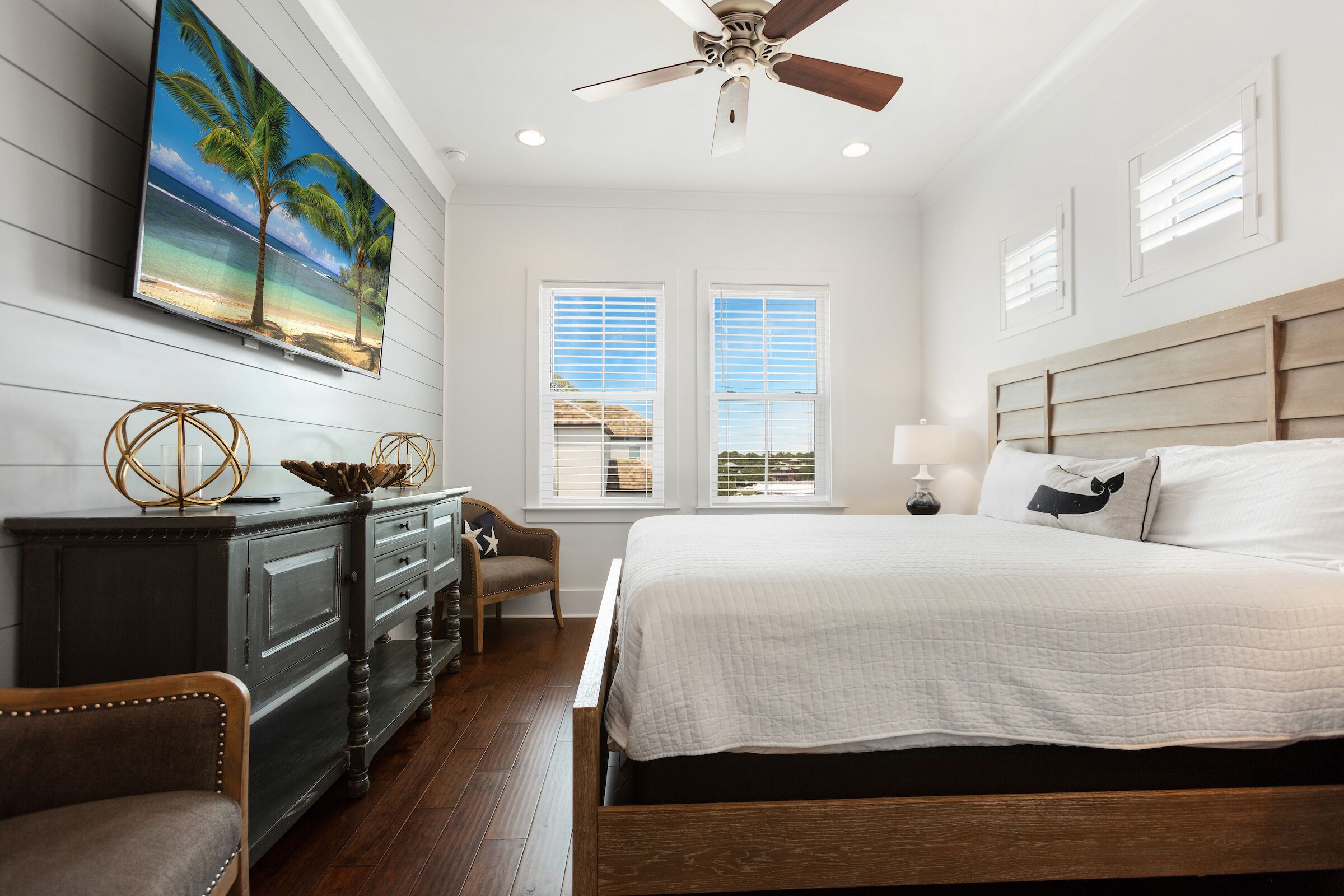 Une chambre à coucher avec un grand lit, un ventilateur de plafond, une commode et un tableau représentant une scène de plage.