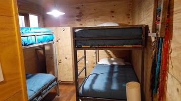 Una habitación pequeña de madera con literas, una cama individual y una mesita con una lámpara.