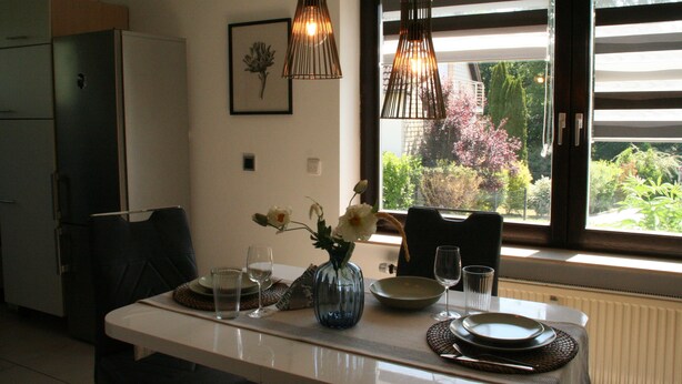 Un comedor con una mesa preparada para dos, un jarrón con flores y una ventana con persianas.