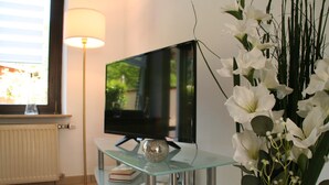 Una sala de estar moderna con un televisor de pantalla plana sobre una mesa de vidrio, una lámpara de pie y un jarrón de flores blancas.