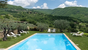 A rectangular swimming pool with a clear blue water surface, surrounded by a grassy area with white lounge chairs. In the background, there are rolling green hills and a partly cloudy sky. - Villa Olivi Pool by Geco Vacation Rentals (Pergine Valdarno)