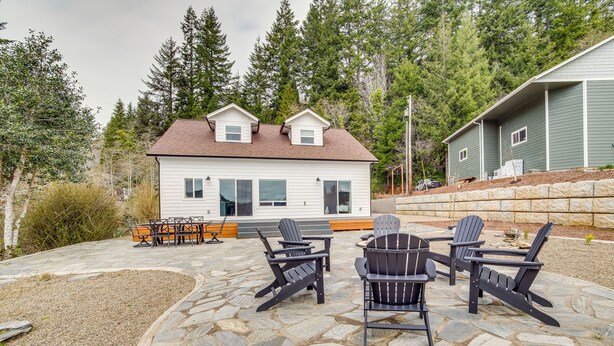 A house with a stone patio, outdoor seating, and a forest backdrop. - Charming Florence Home w/ Fire Pit & River Views! (Florence)