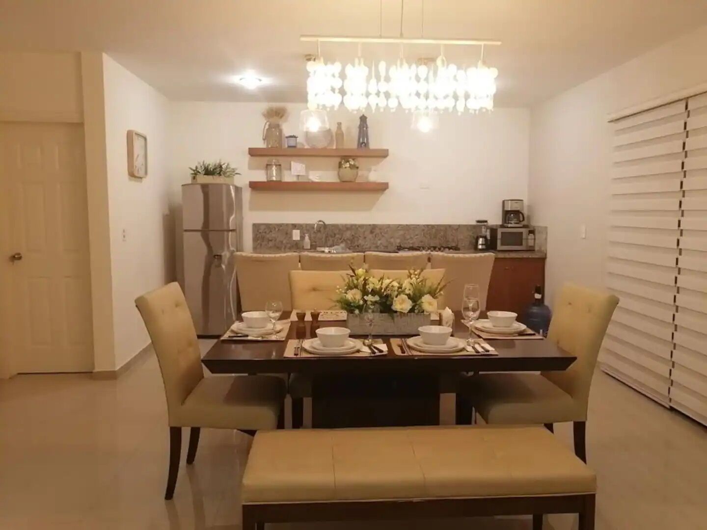 Un coin repas avec une table dressée pour quatre personnes, un lustre au-dessus et une cuisine en arrière-plan.