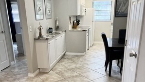 Una cocina con armarios blancos, una zona de comedor con mesa y sillas negras, y un piso con baldosas.
