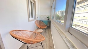 Un balcone con due sedie in vimini, un tavolino e vista su edifici attraverso ampie finestre.