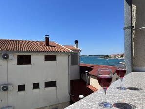 A balcony with a view of the sea, a building with a tiled roof, and two glasses of red wine.