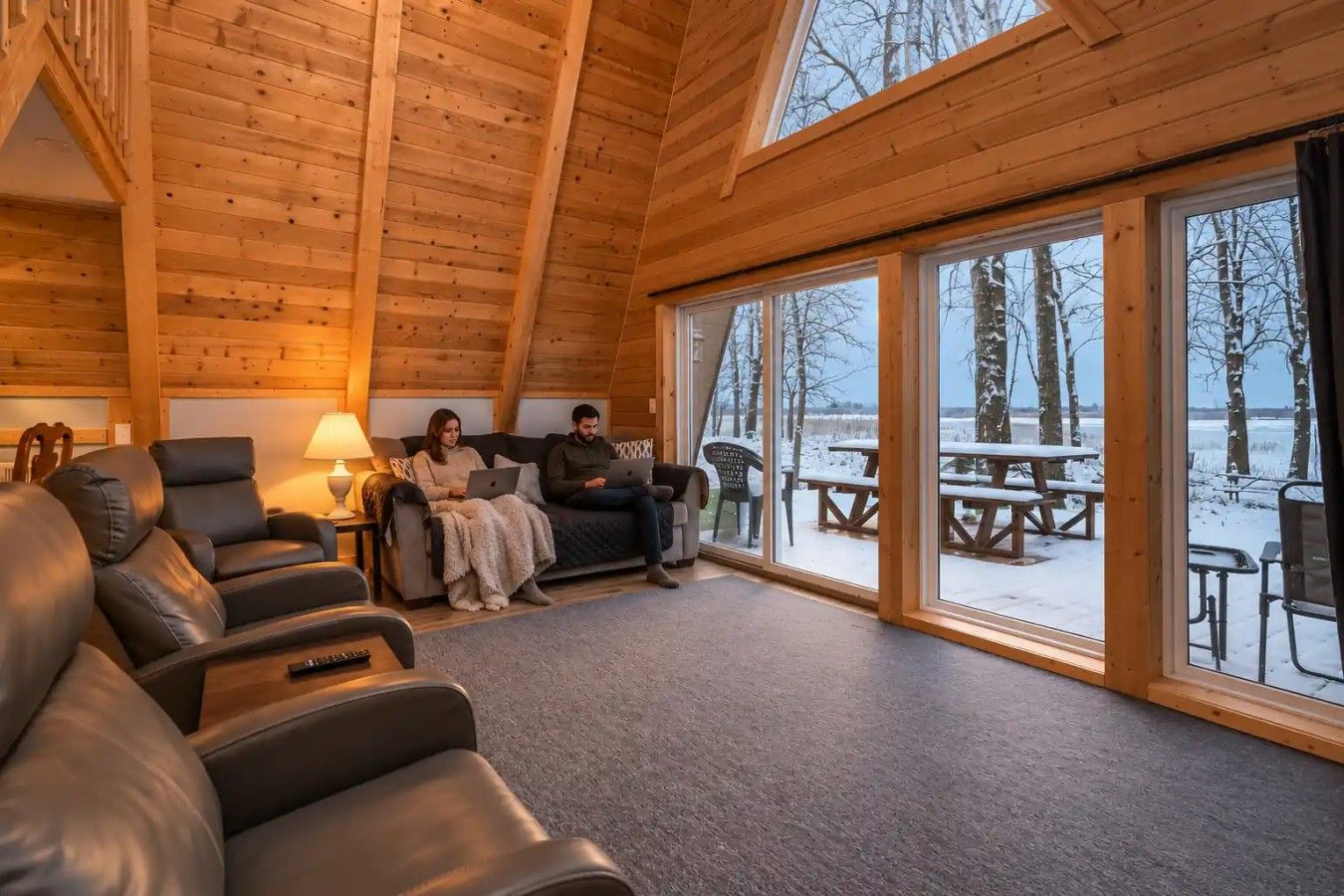 A wooden cabin interior with a sofa, chairs, and a view of a snowy outdoor area with picnic tables.