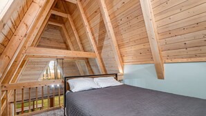 A bedroom with a sloped ceiling, wooden beams, a bed with a grey quilt, and a view of the outdoors. - Stunning Sunrise Shore (Au Gres)