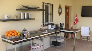 Une cuisine avec un long comptoir, une machine à café, une bouilloire et un saladier rempli de fruits.