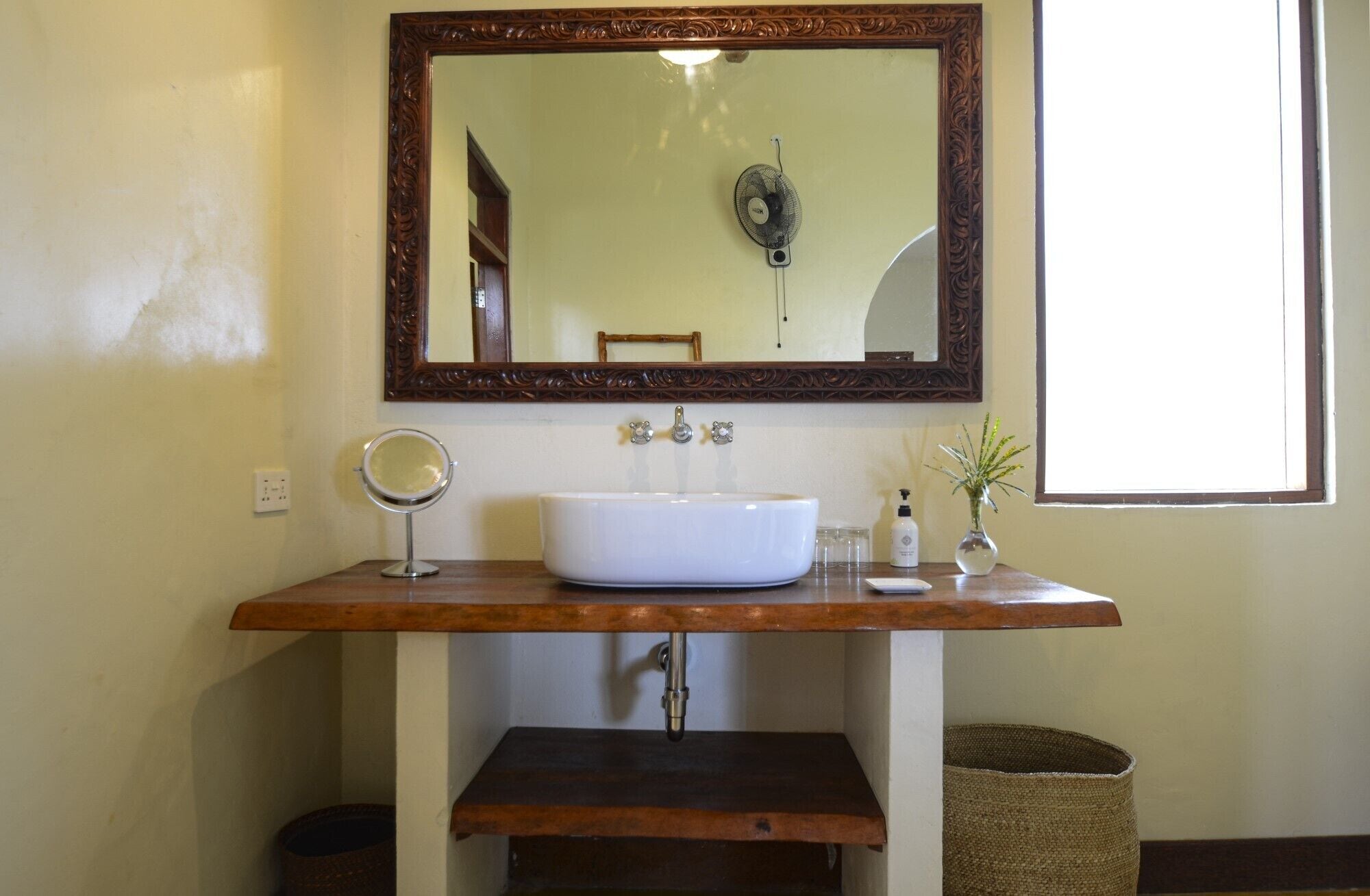 Un baño con tocador de madera, un lavamanos blanco, un espejo redondo, un ventilador y una ventana.