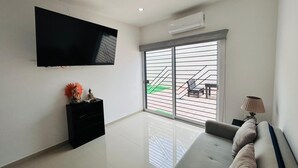 A modern living room with a flat-screen TV, a sofa, a black entertainment unit, and a large sliding glass door leading to an outdoor patio. - Napoles (Mazatlán)