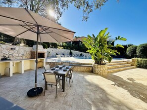 Terrassa exterior amb mur de pedra, una barbacoa, una piscina i zona de dinar amb cadires i taula.