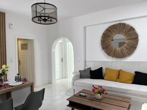 A modern living room with a white sofa, a round mirror, and a dining area with a table and chairs. - 3 Bedroom House included car rental-Heated Jacuzzi (santorini)