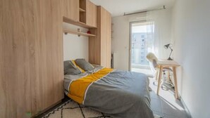 Une chambre à coucher avec un lit, un bureau et une chaise. Il y a une fenêtre qui donne sur des immeubles.