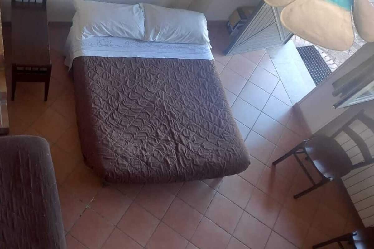 Une pièce avec un lit, une petite table et une chaise. On aperçoit un escalier au fond.