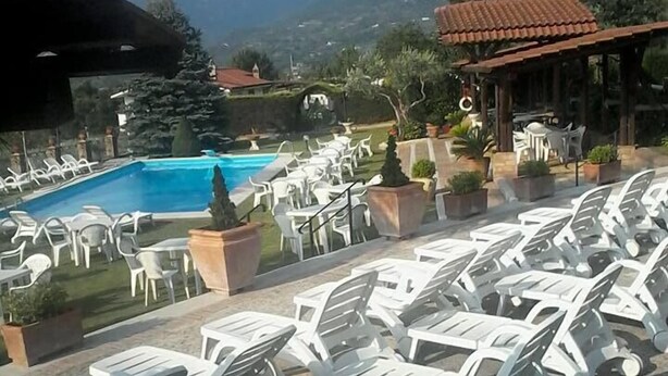 Zona junto a la piscina con sillas y mesas de plástico blanco, una piscina azul cristalina y un paisaje montañoso al fondo.