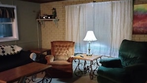 A cozy room with a wooden ceiling, a green armchair, a tufted armchair, a small table with a lamp, and a window with curtains.