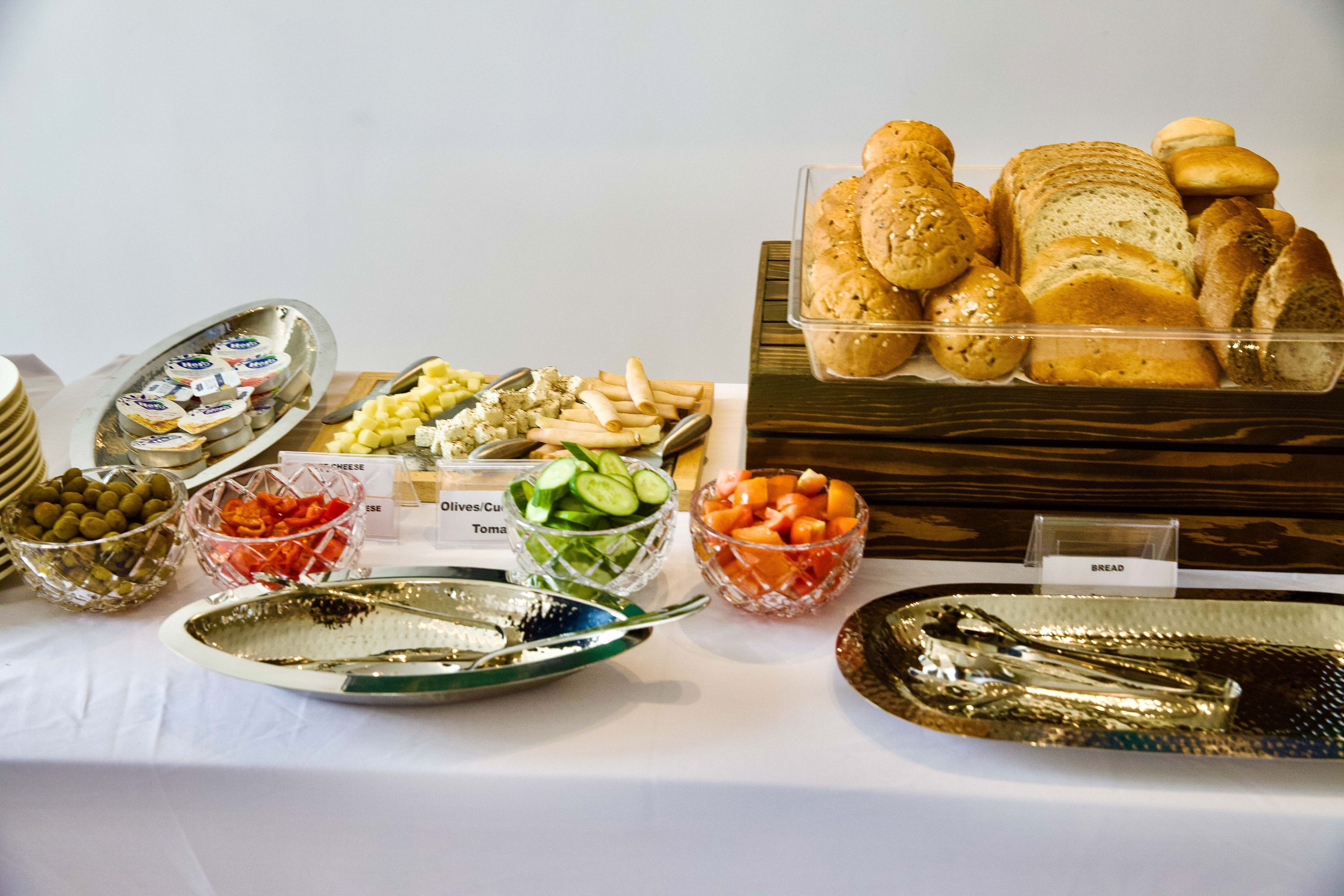Una mesa de buffet con diversos alimentos, entre ellos pan, queso, aceitunas, pepinos, tomates y un plato de pescado.