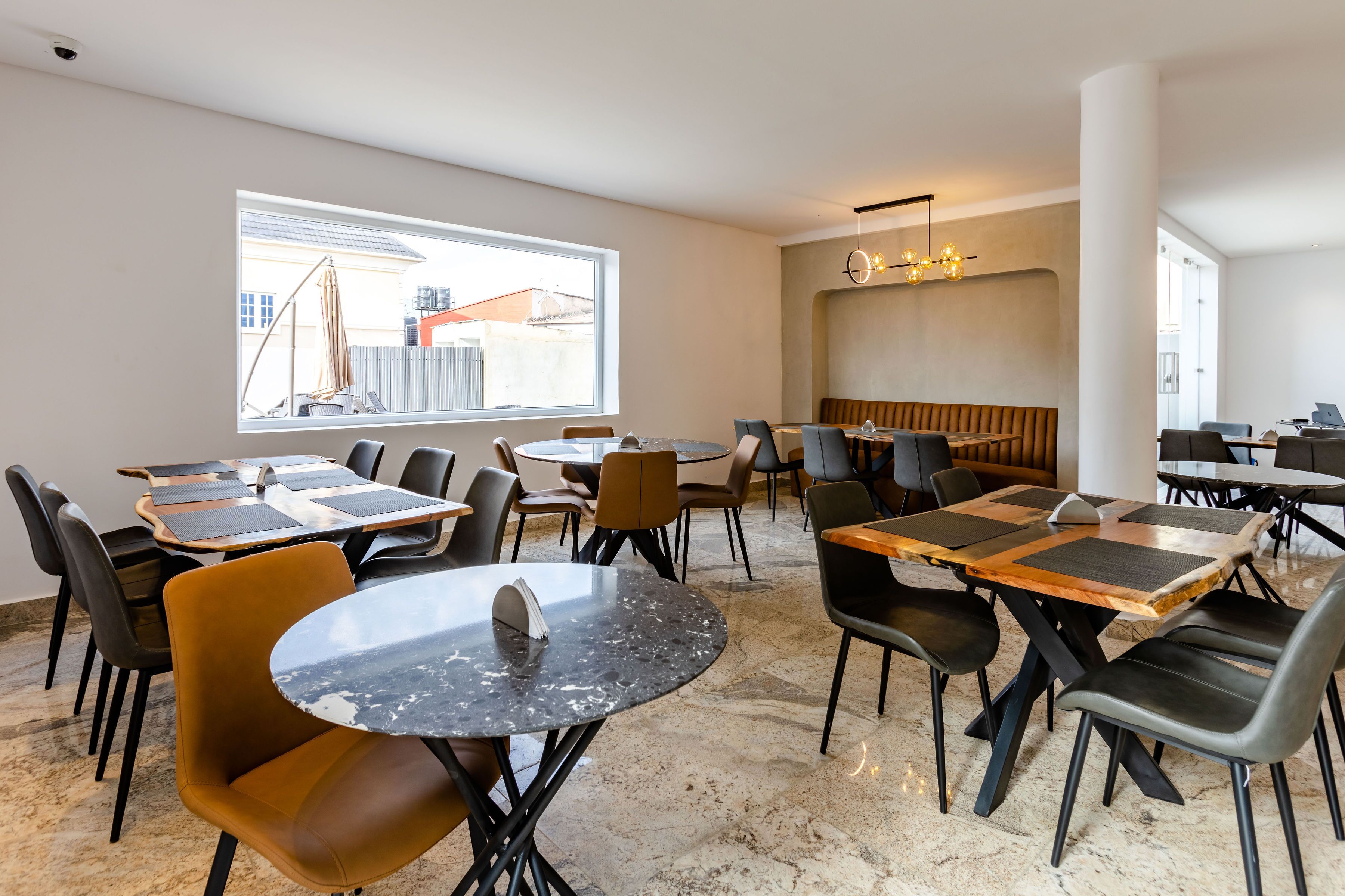 A modern dining area with multiple tables and chairs, a large window with a view of buildings, and a contemporary light fixture.
