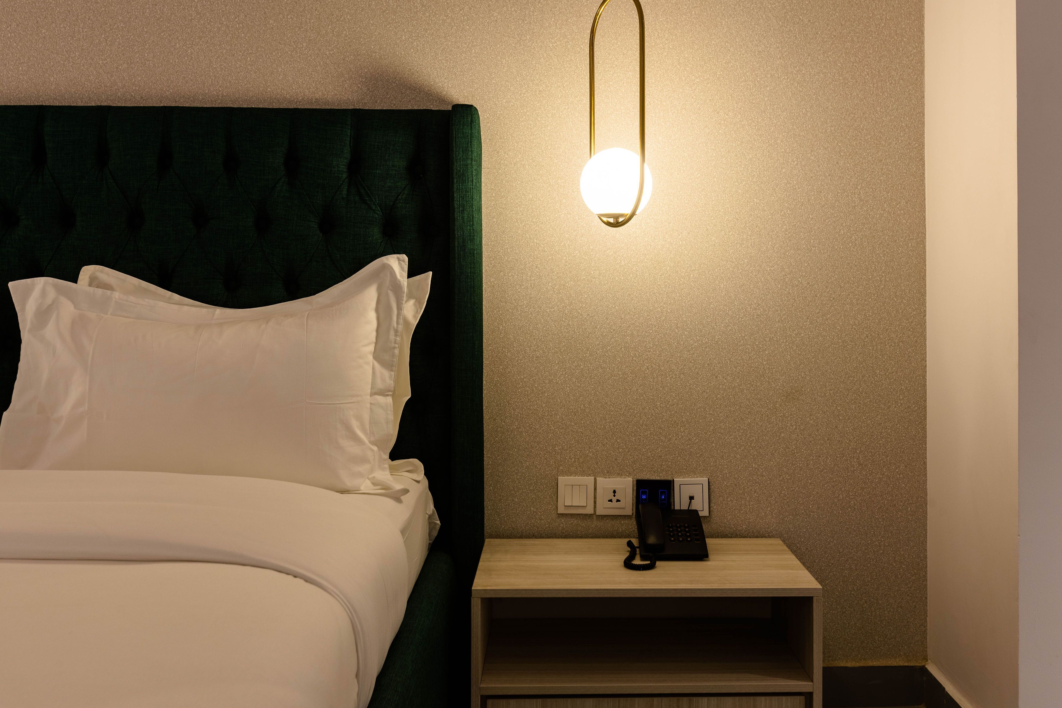 Un letto rifatto con lenzuola bianche e una testiera imbottita verde. Si vede un comodino con un telefono e una lampada.