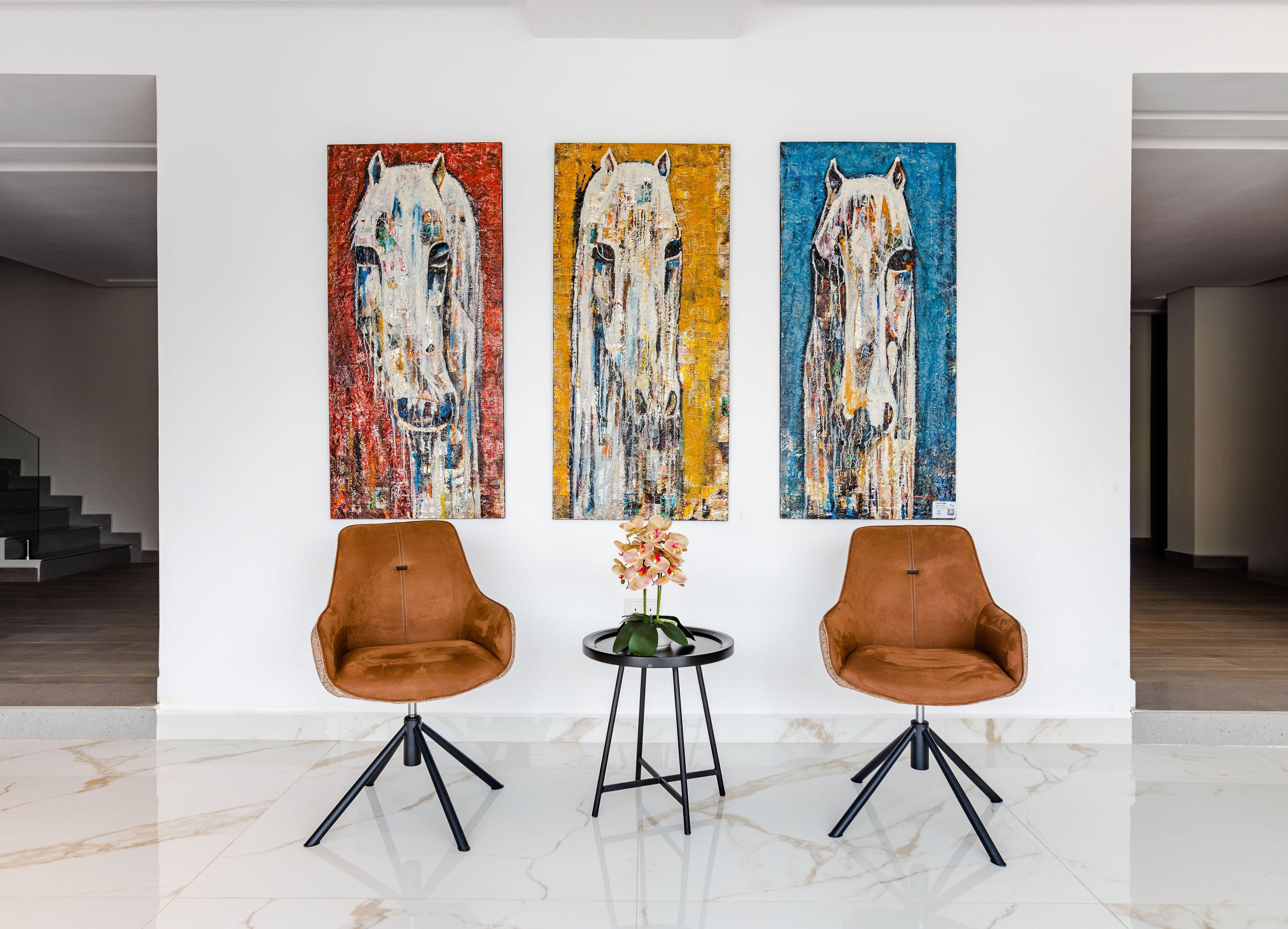 Un interior moderno con tres cuadros abstractos de caballos en la pared, dos sillones de cuero marrón y una pequeña mesa con un jarrón de flores.