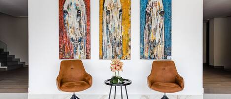 Un interior moderno con tres cuadros abstractos de caballos en la pared, dos sillones de cuero marrón y una pequeña mesa con un jarrón de flores.